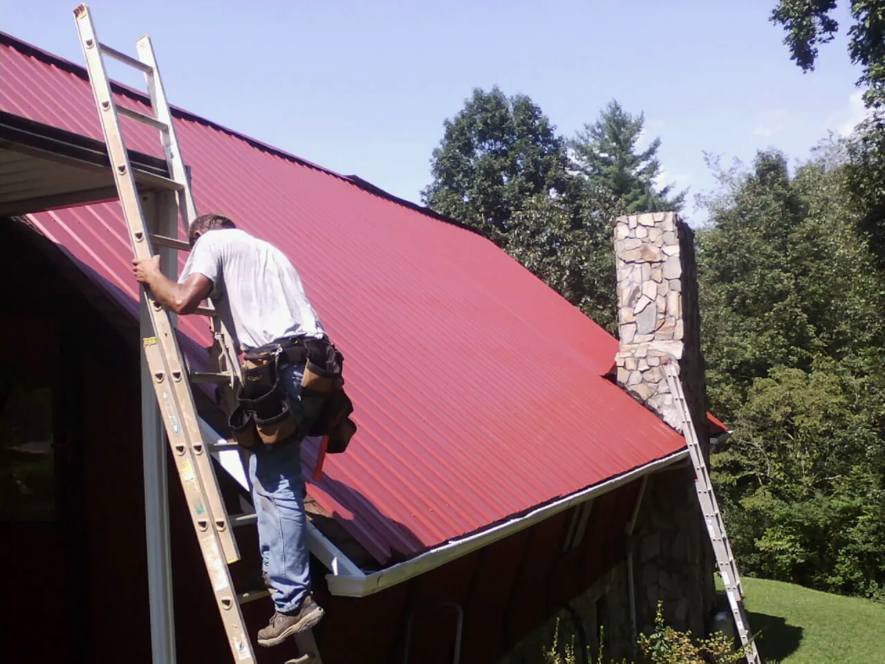 Roofer on a ladder beside a red metal roof and stone chimney — Soffit & Fascia Repair by Roofing Brothers in Mount Prospect